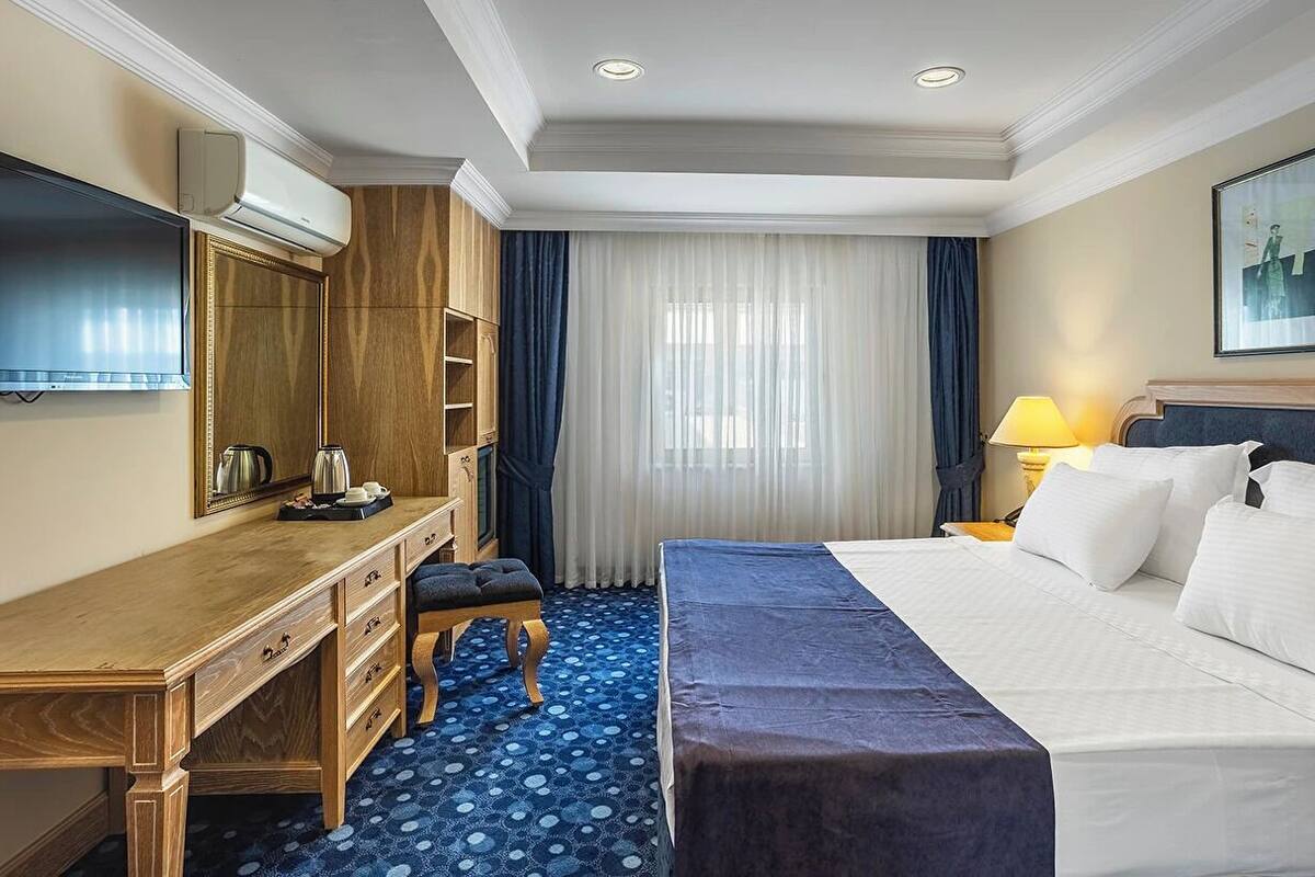 Una habitación de hotel con cama, escritorio, televisor, cafetera y ventana con cortinas.
