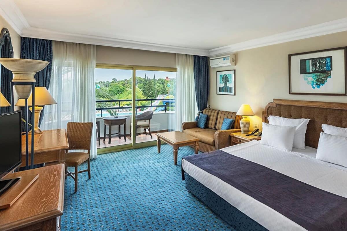 Habitación de hotel con una cama grande, un sofá, un escritorio con lámpara, una silla y un balcón con vistas a zonas verdes.