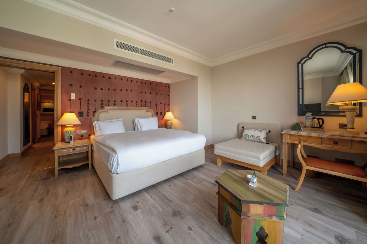 Habitación de hotel con una cama grande, dos mesitas de noche con lámparas, un escritorio de madera, una silla, una mesita auxiliar colorida y un espejo en la pared.
