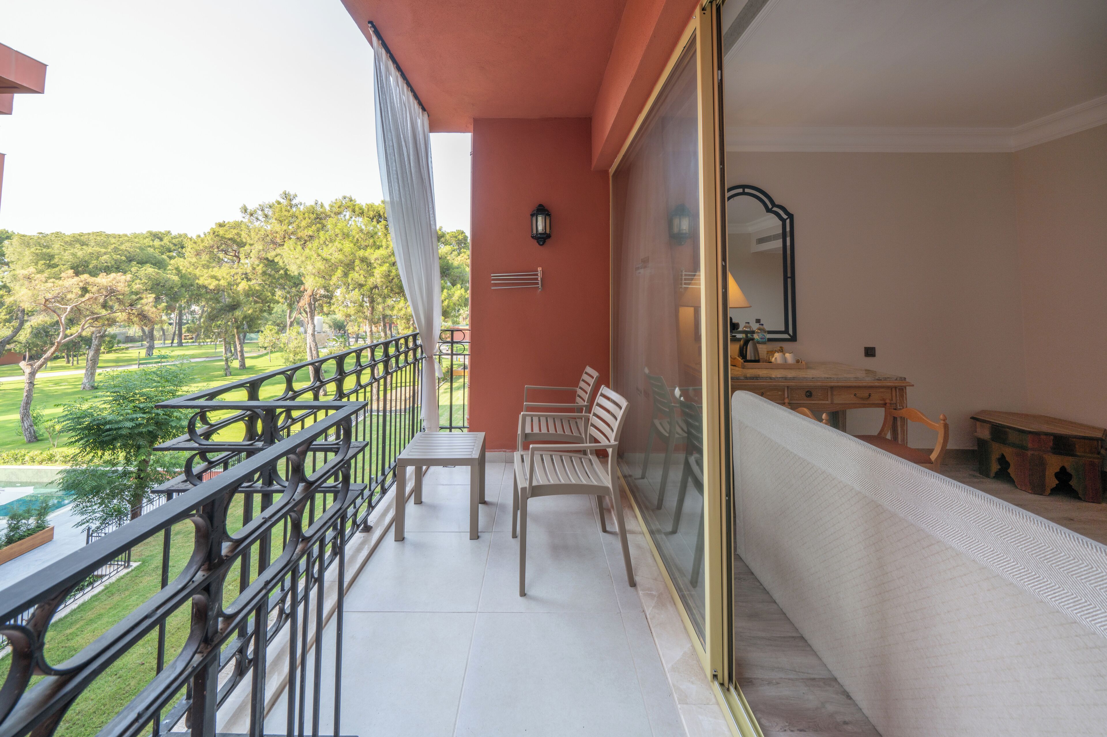 A balcony with a table and chairs, a view of a park, and a sliding glass door leading to a room with a mirror and a small table.