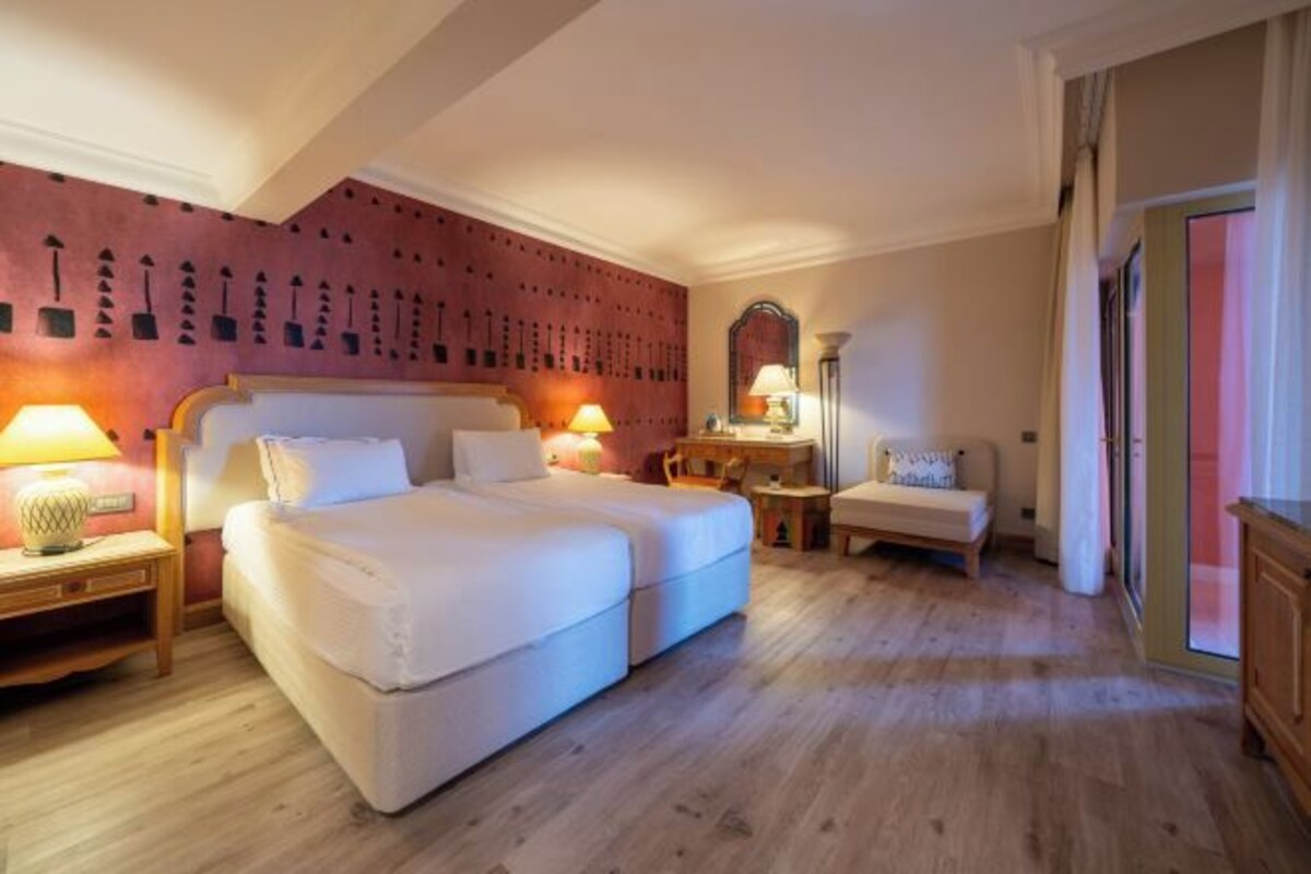 A hotel room with a large bed, two bedside tables with lamps, a small chair, a mirror, and a wooden floor.