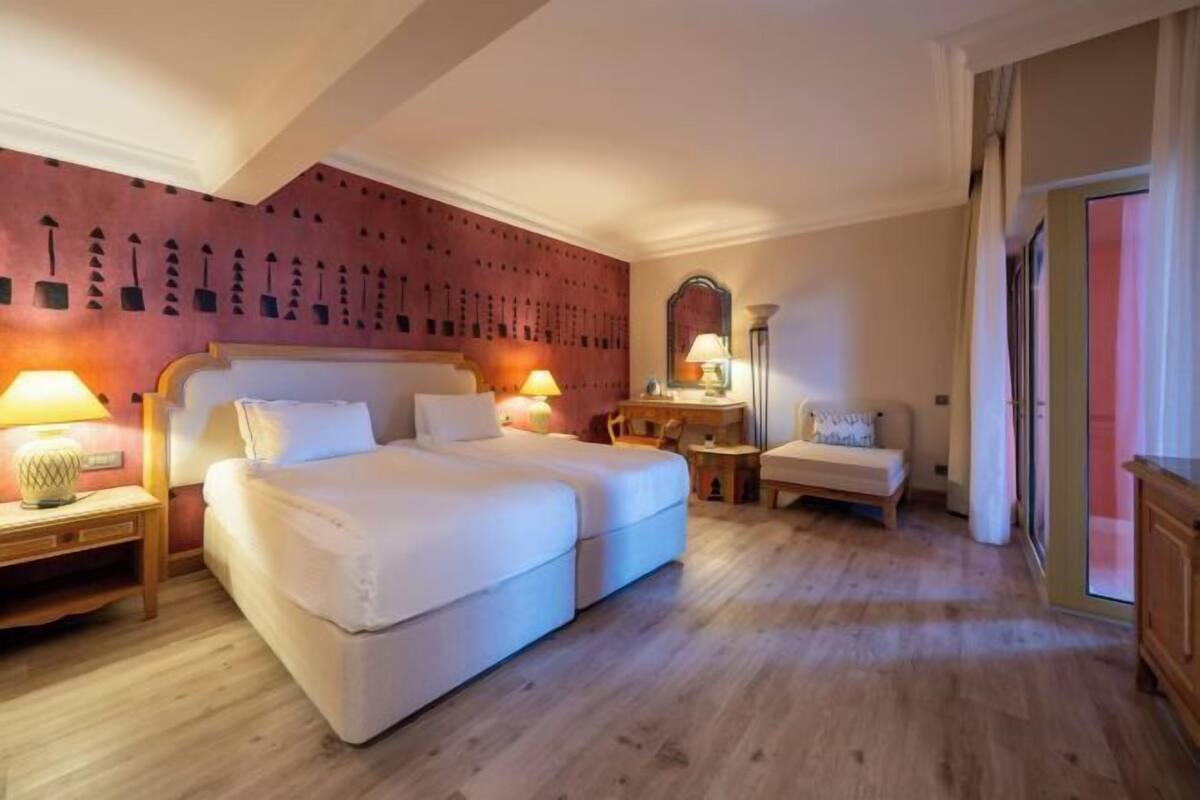 Una habitación de hotel con una cama grande, dos mesitas de noche con lámparas, una silla pequeña, un espejo y suelo de madera.