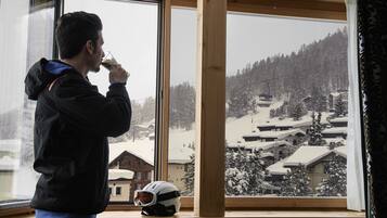 Una persona con chaqueta negra bebiendo de un vaso, con un casco en el alféizar de la ventana y una vista nevada de un pueblo afuera.