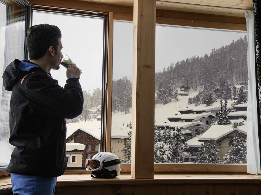 Una persona con chaqueta negra bebiendo de un vaso, con un casco en el alféizar de la ventana y una vista nevada de un pueblo afuera.