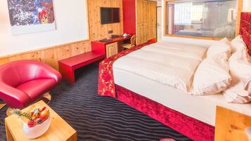 A hotel room with a bed, a desk, a red chair, a small table with fruit, and a mirror.