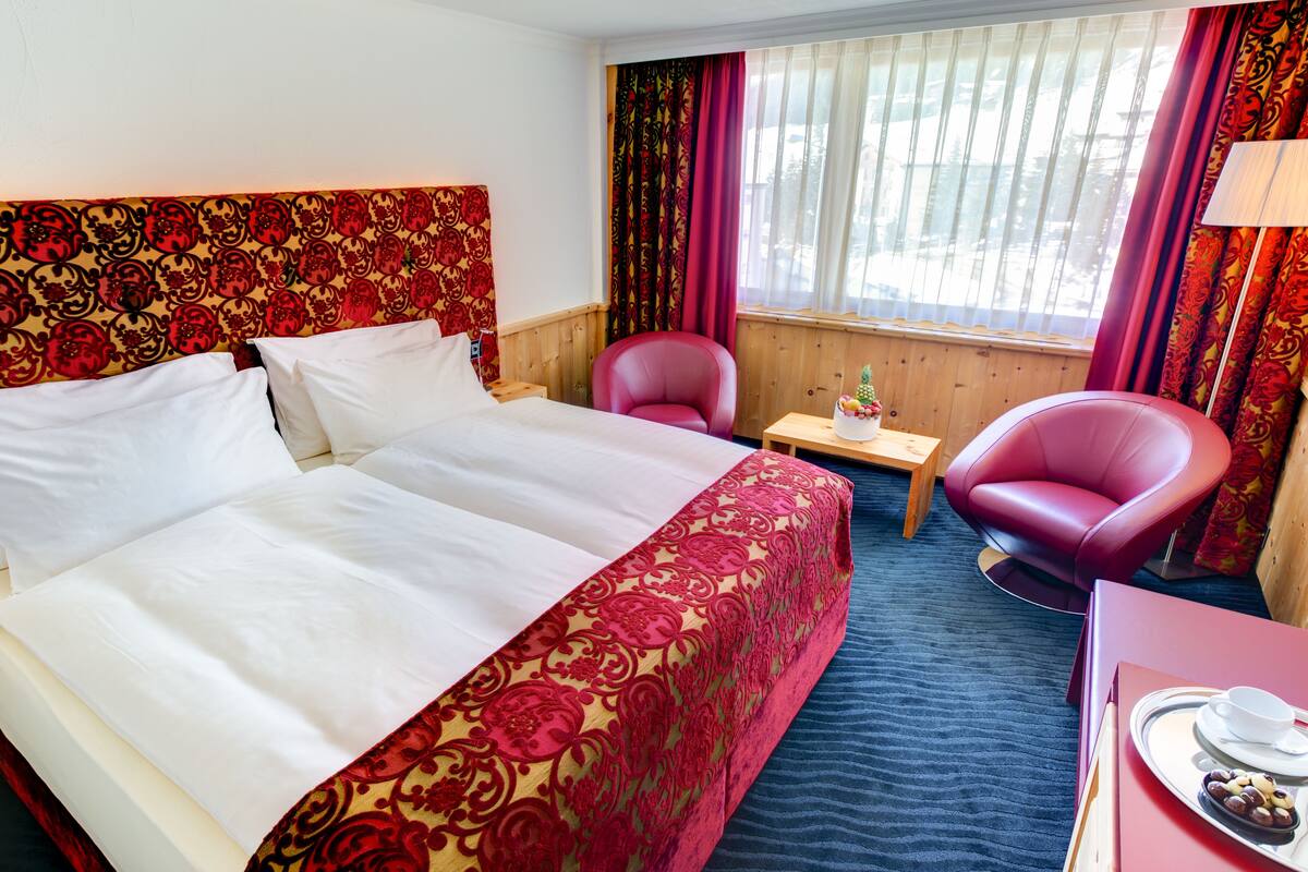 Ein Hotelzimmer mit einem großen Bett, zwei Stühlen, einem kleinen Tisch und einem Fenster mit Vorhängen.