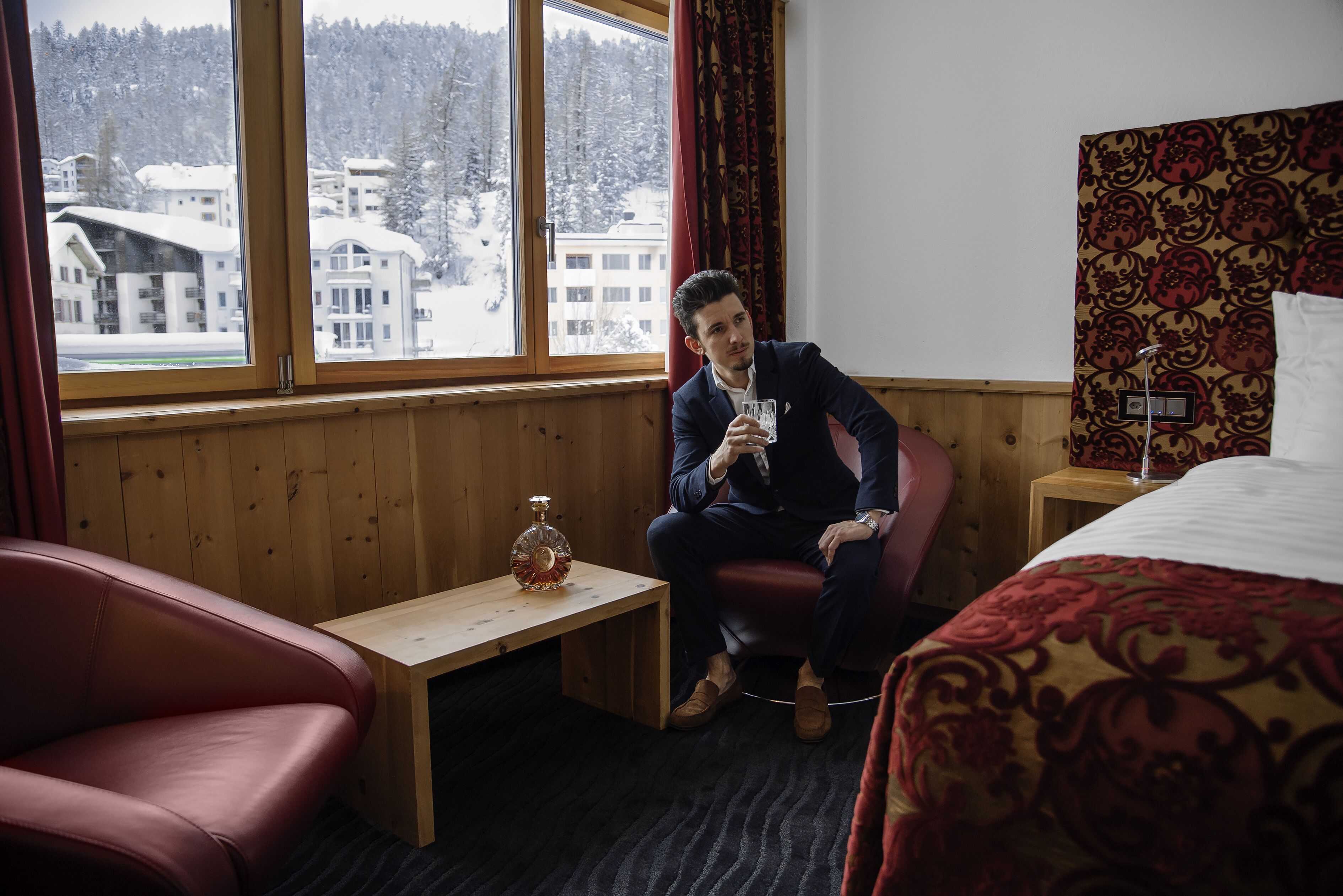 Ein Hotelzimmer mit einer holzverkleideten Wand, einem großen Fenster mit Blick auf schneebedeckte Gebäude, einem kleinen Holztisch mit einer Flasche und einem roten Ledersessel.