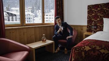Ein Hotelzimmer mit einer holzverkleideten Wand, einem großen Fenster mit Blick auf schneebedeckte Gebäude, einem kleinen Holztisch mit einer Flasche und einem roten Ledersessel.