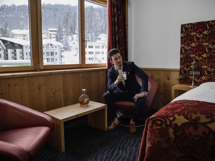 A hotel room with a wooden paneled wall, a large window with a view of snow-covered buildings, a small wooden table with a bottle, and a red leather chair.