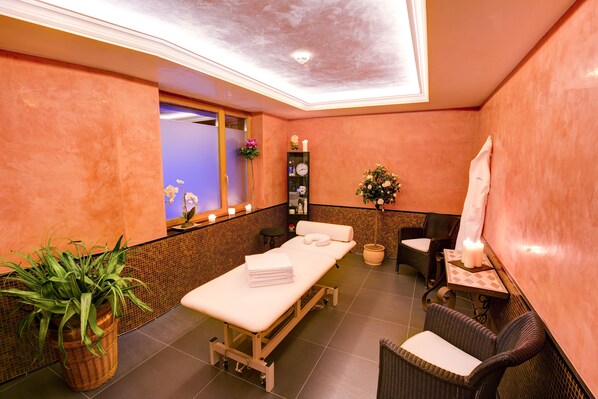 A spa room with a massage table, chairs, a small table with candles, and a potted plant. - Hotel San Gian (St. Moritz)
