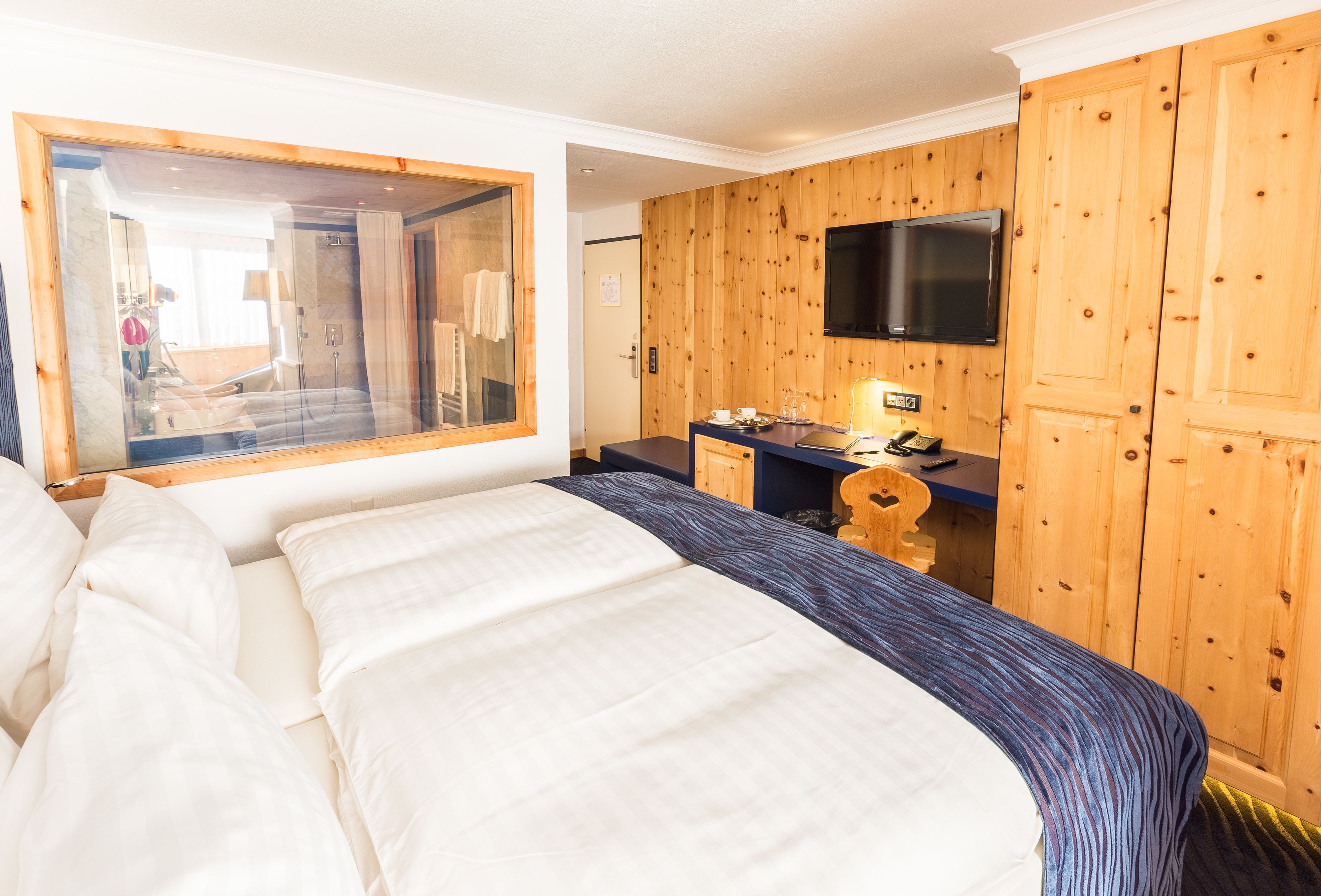 A hotel room with a large bed, a wooden wardrobe, a desk with a chair, and a mounted TV.