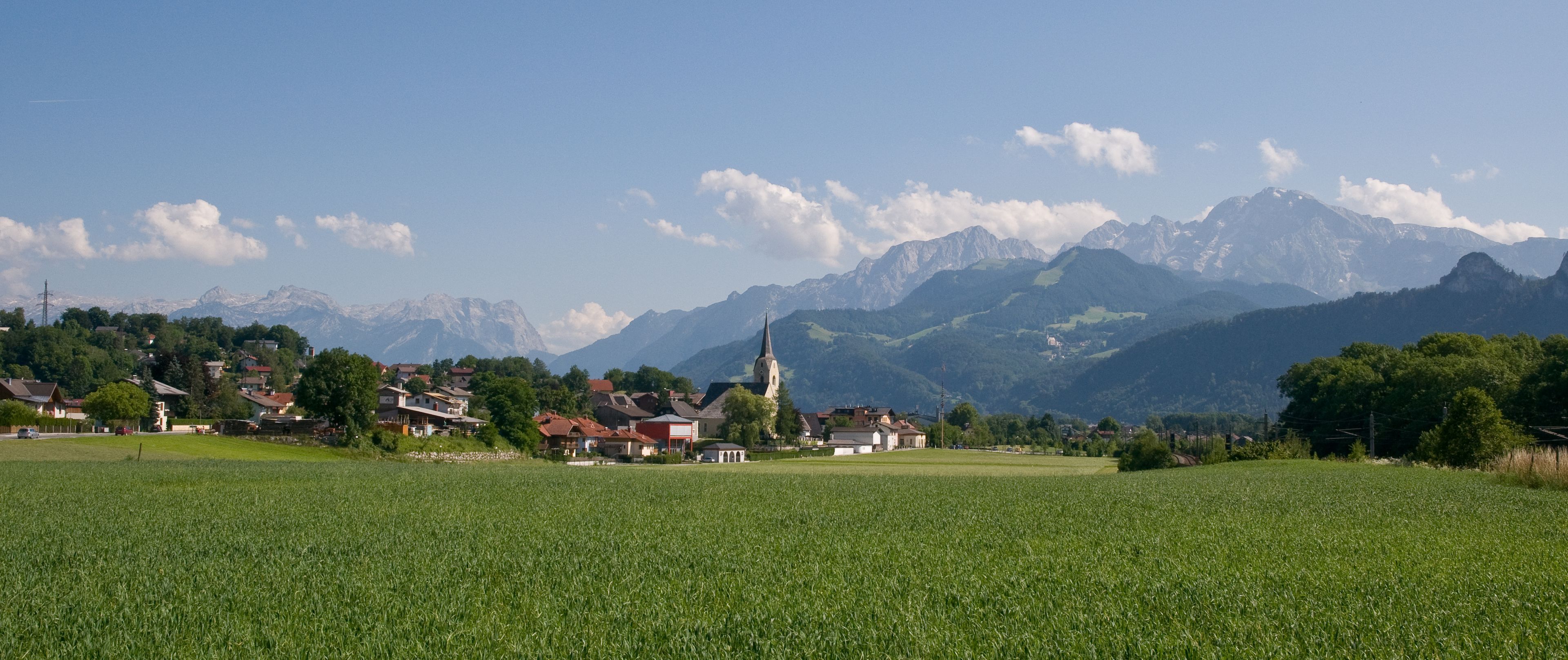 Ein ländliches Dorf mit Kirche, Häusern und einem Feld im Vordergrund, mit Bergen im Hintergrund.