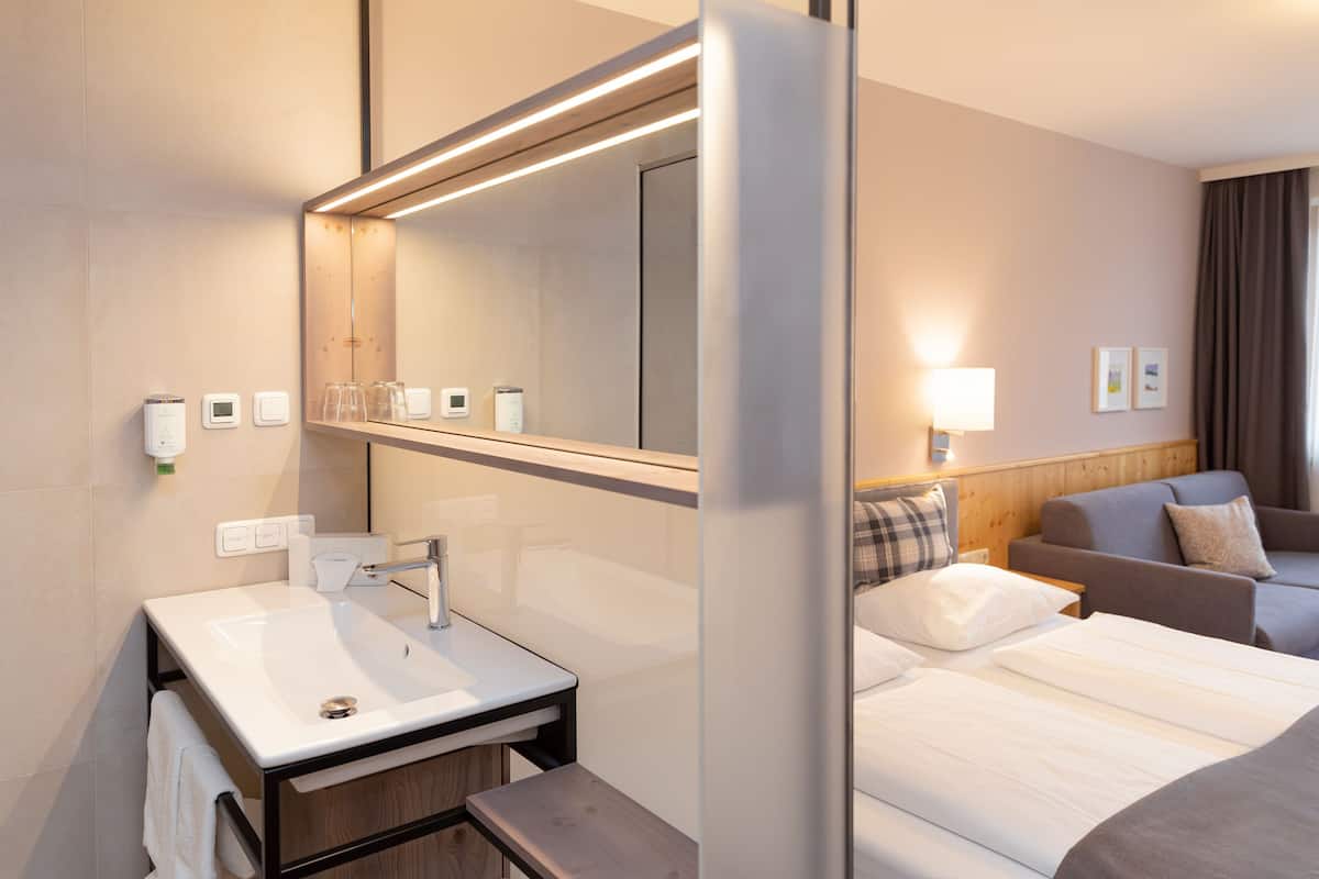 A hotel room with a large mirror, a sink, a bed, a sofa, and a wall-mounted lamp.