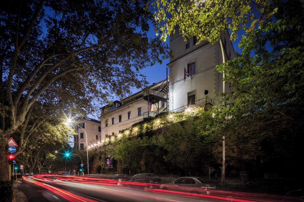 A street at dusk with a building on the right, trees, and light trails from moving vehicles.