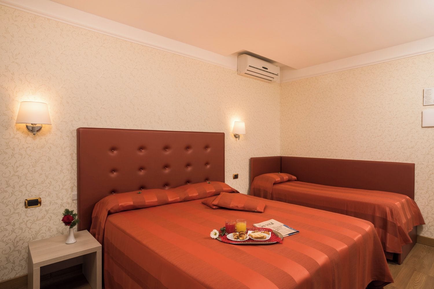 A hotel room with two beds, a nightstand, a lamp, and a small table with breakfast items.