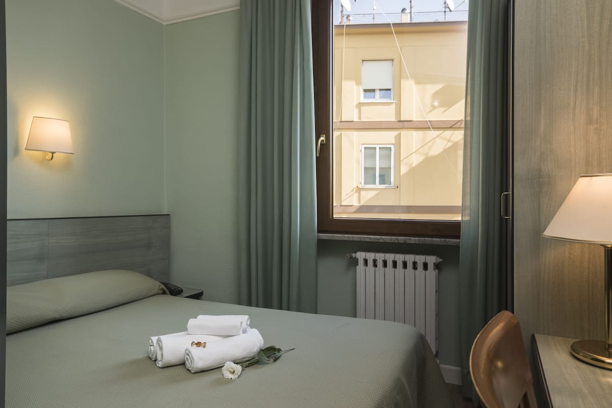 A hotel room with a bed, a nightstand, a lamp, a window with curtains, and a radiator.