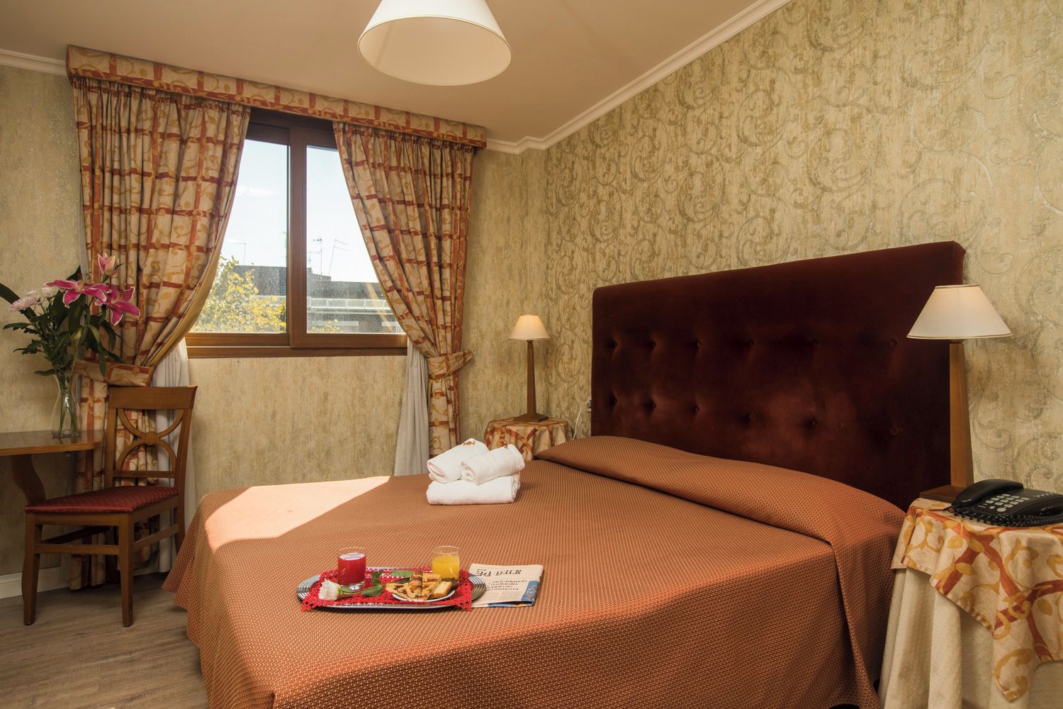 Ein Hotelzimmer mit Bett, zwei Nachttischlampen, einem Tisch mit Frühstücksbrettchen, einem Stuhl und einem Fenster mit Vorhängen.