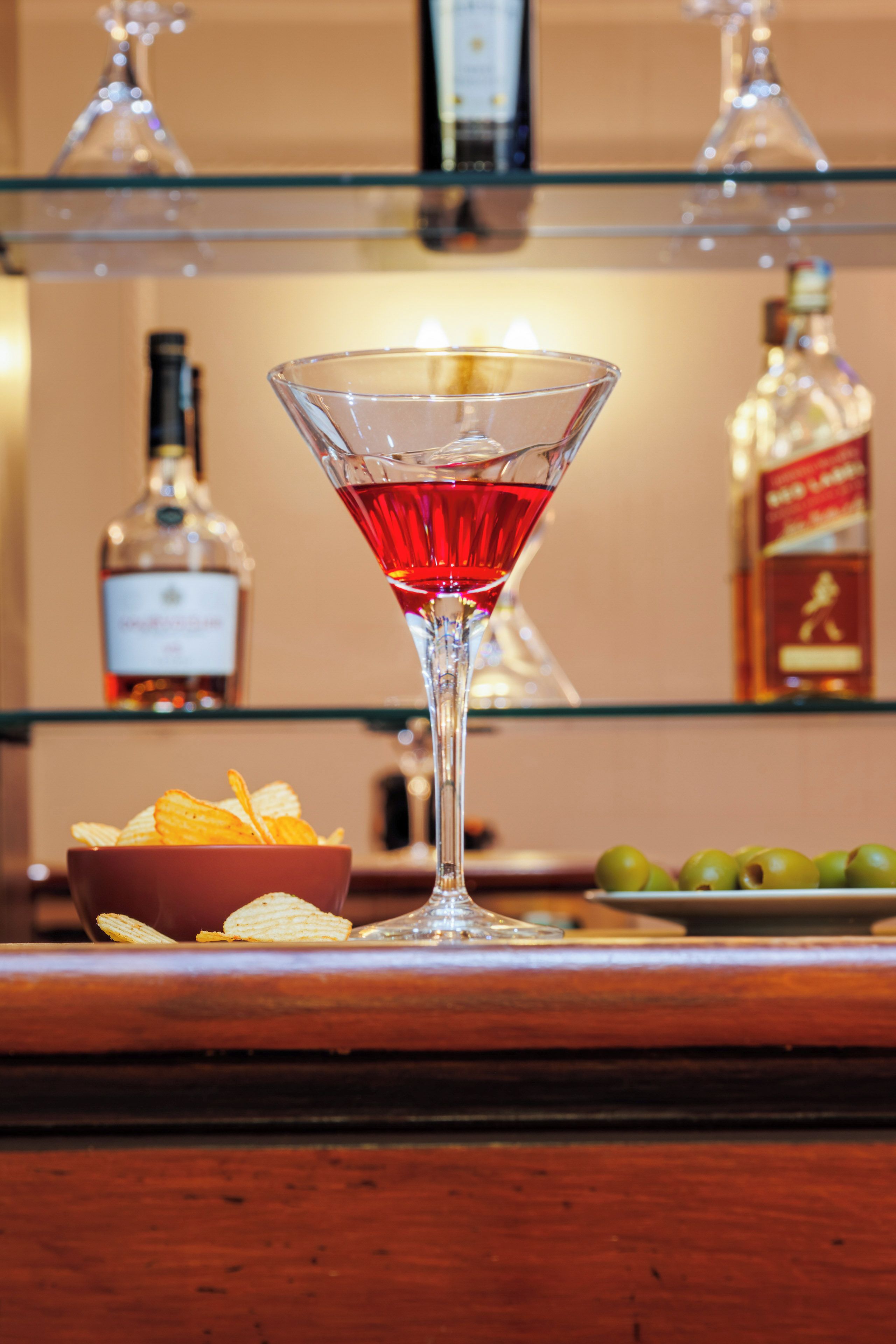 Un bar con una copa de martini llena de una bebida roja, un cuenco de papas fritas y aceitunas sobre la barra.