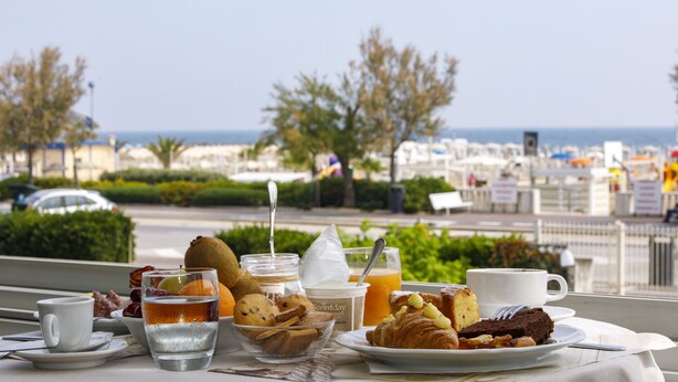 A breakfast table with croissants, cookies, and a fruit bowl, overlooking a marina with boats. - Hotel Derby (Rimini)