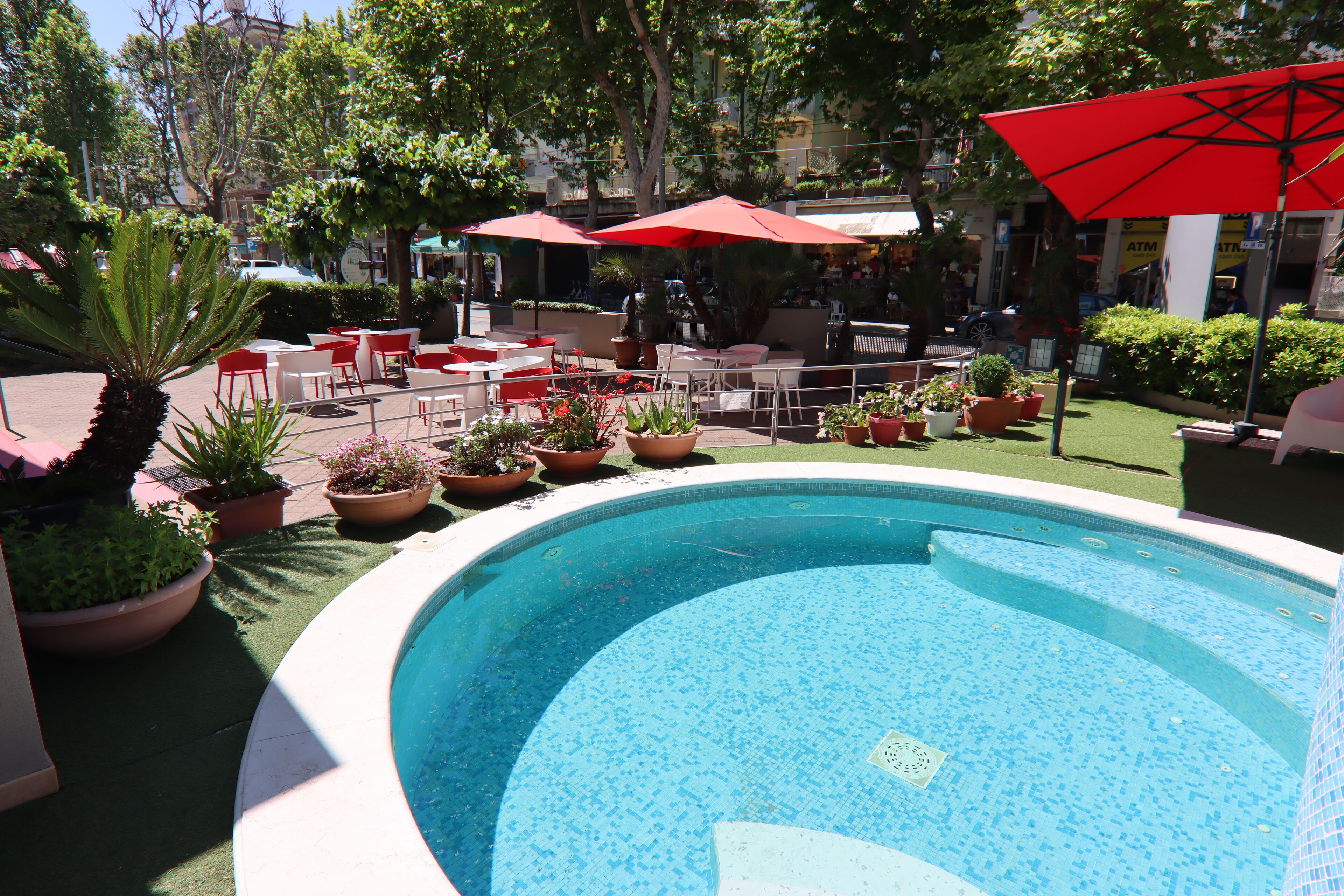 A swimming pool surrounded by outdoor seating with red umbrellas, potted plants, and a green lawn.