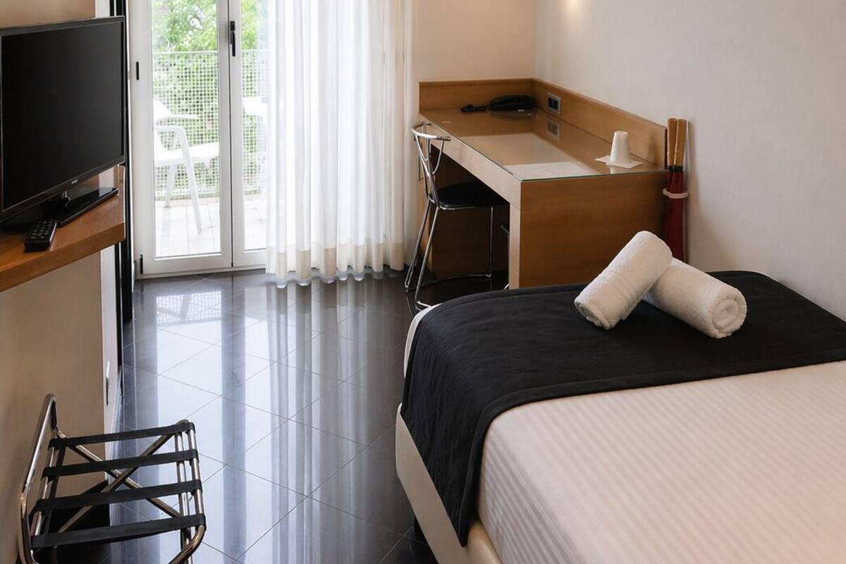 A hotel room with a bed, a desk, a TV, a bathroom, and a view through a glass door.