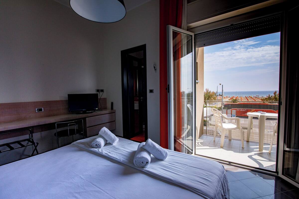A hotel room with a bed, a TV, a desk, and a balcony with a view of the sea.