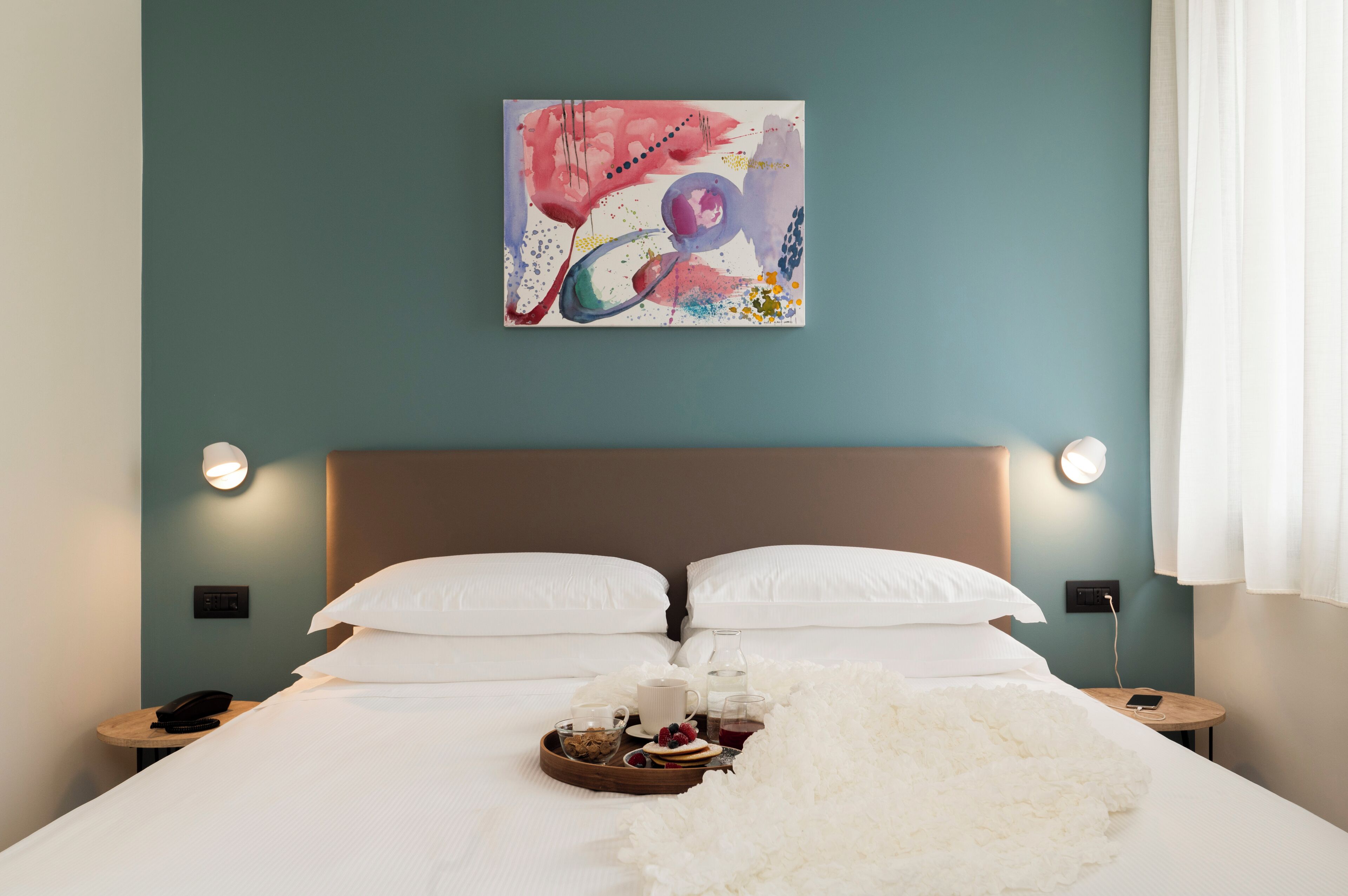 A hotel room with a bed, white pillows, a bedside table with a phone, a tray with breakfast items, and a painting on the wall.