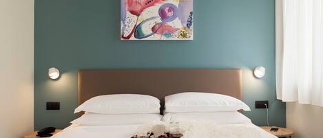 A hotel room with a bed, white pillows, a bedside table with a phone, a tray with breakfast items, and a painting on the wall.