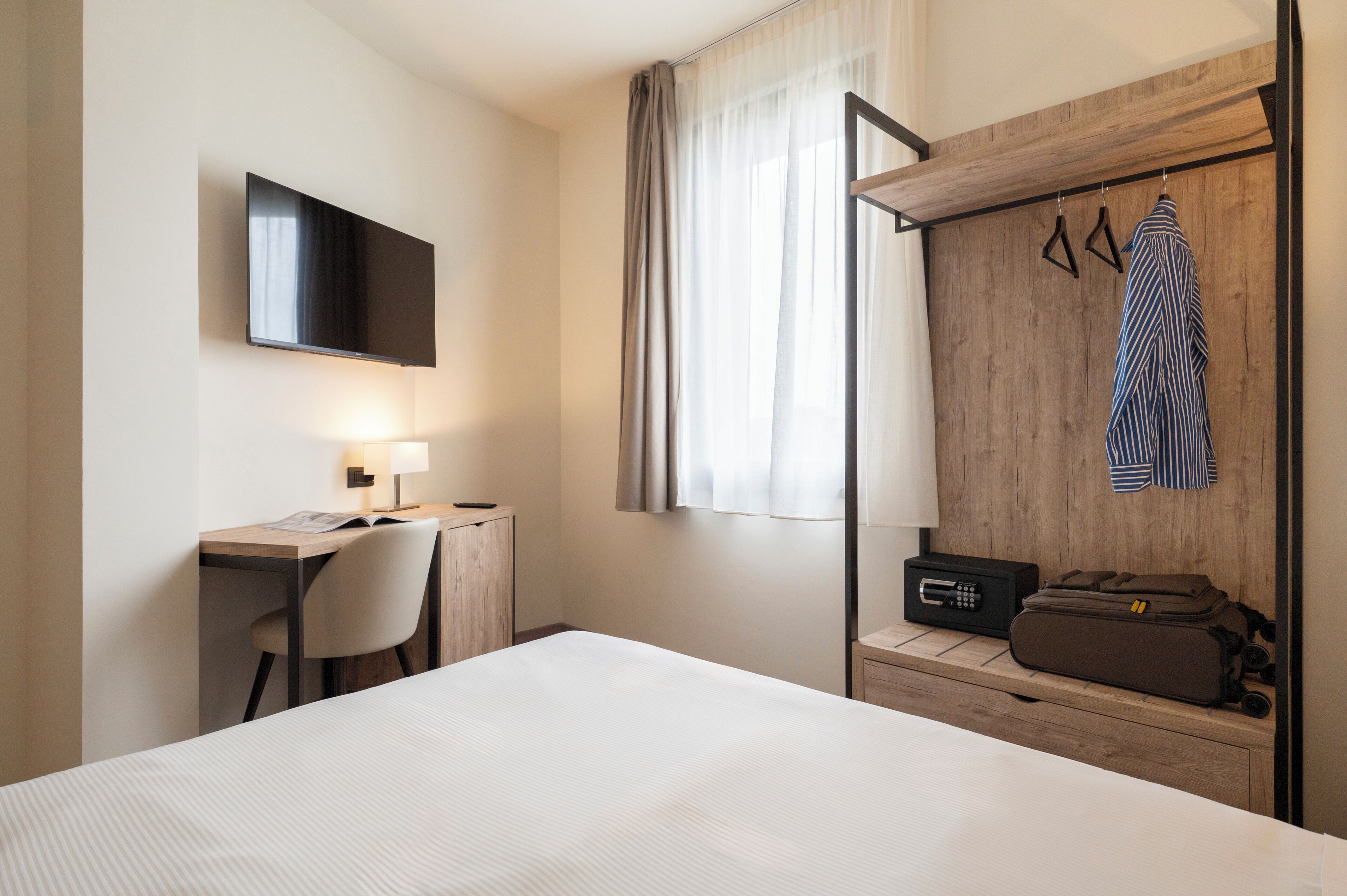 A hotel room with a bed, desk, chair, TV, wardrobe, and a suitcase.