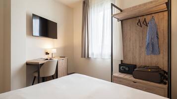 A hotel room with a bed, desk, chair, TV, wardrobe, and a suitcase.
