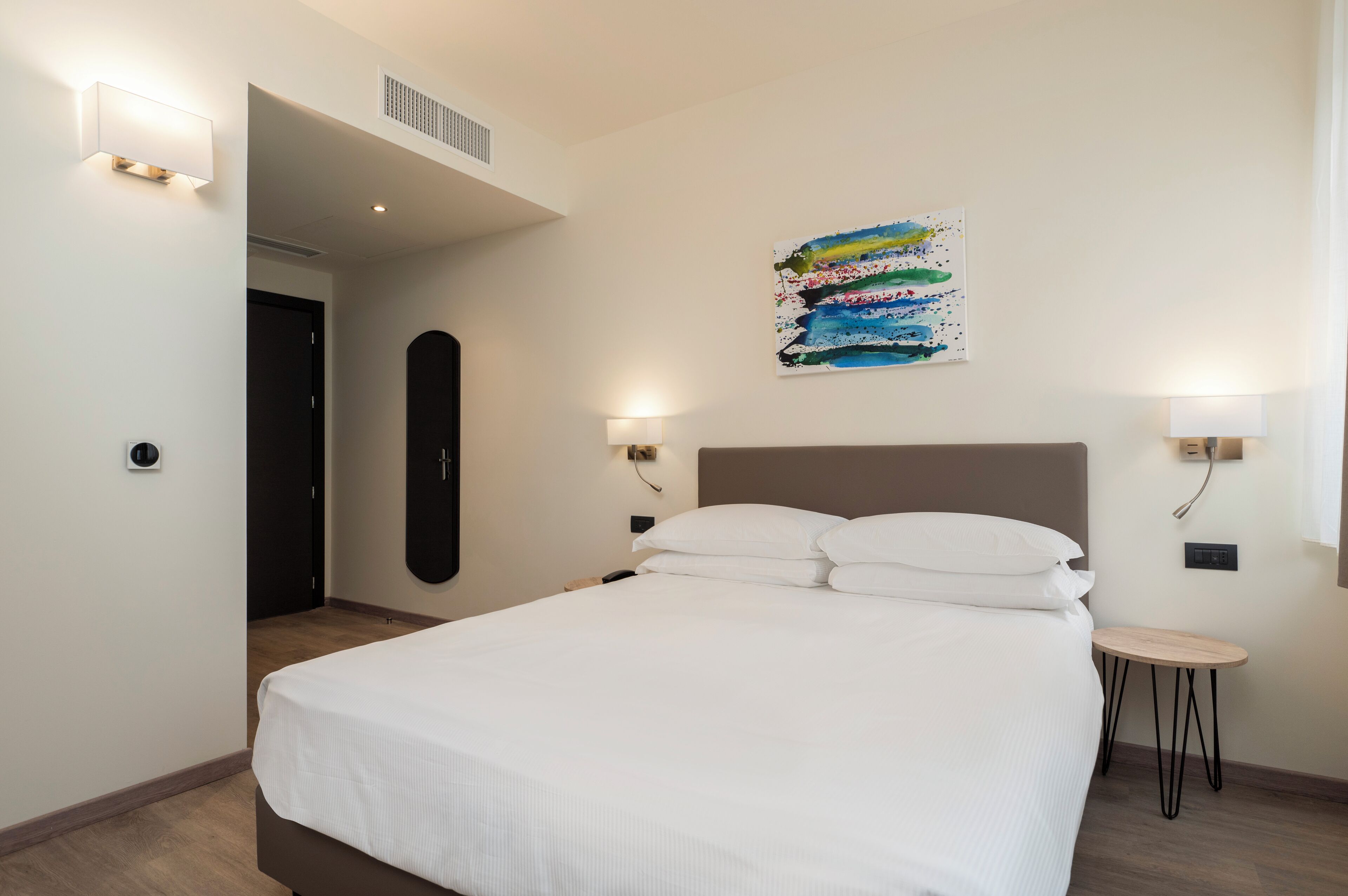 A modern hotel room with a large bed, a small round table, and two wall-mounted lamps.