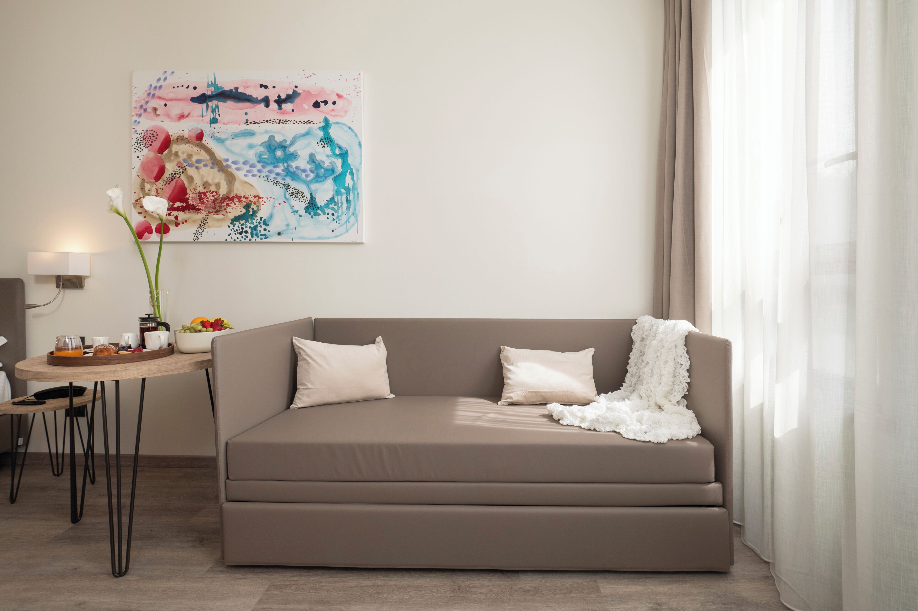 A modern living room with a beige sofa, a small table with a vase of flowers, and a colorful abstract painting on the wall.