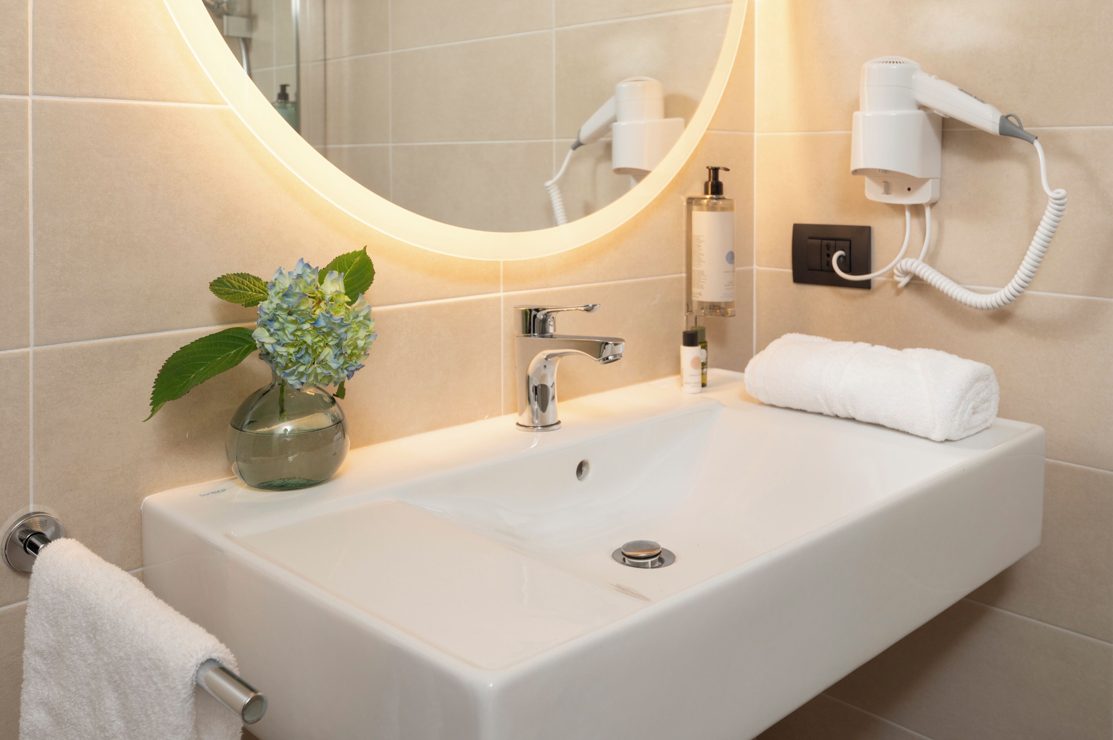 A bathroom with a white sink, a mirror, a hairdryer, and a vase with flowers.