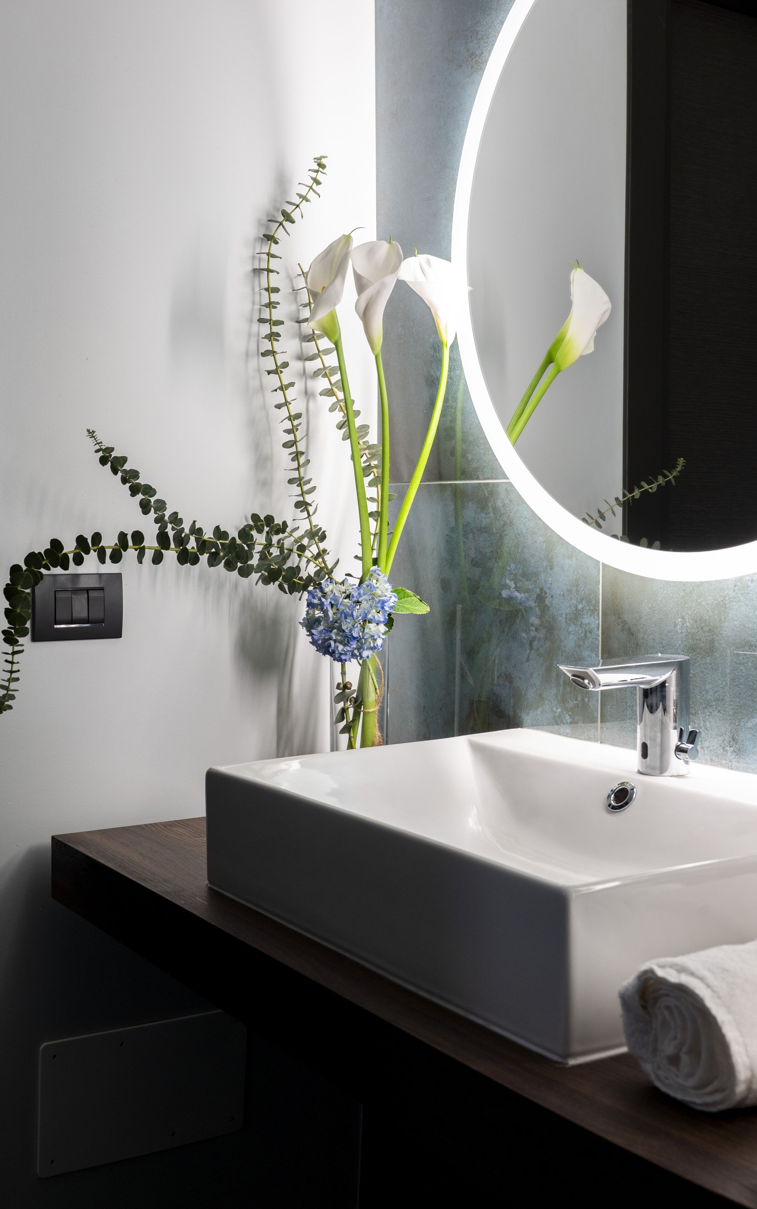 Un baño moderno con un lavamanos blanco, un espejo con luz circular y un florero con flores.