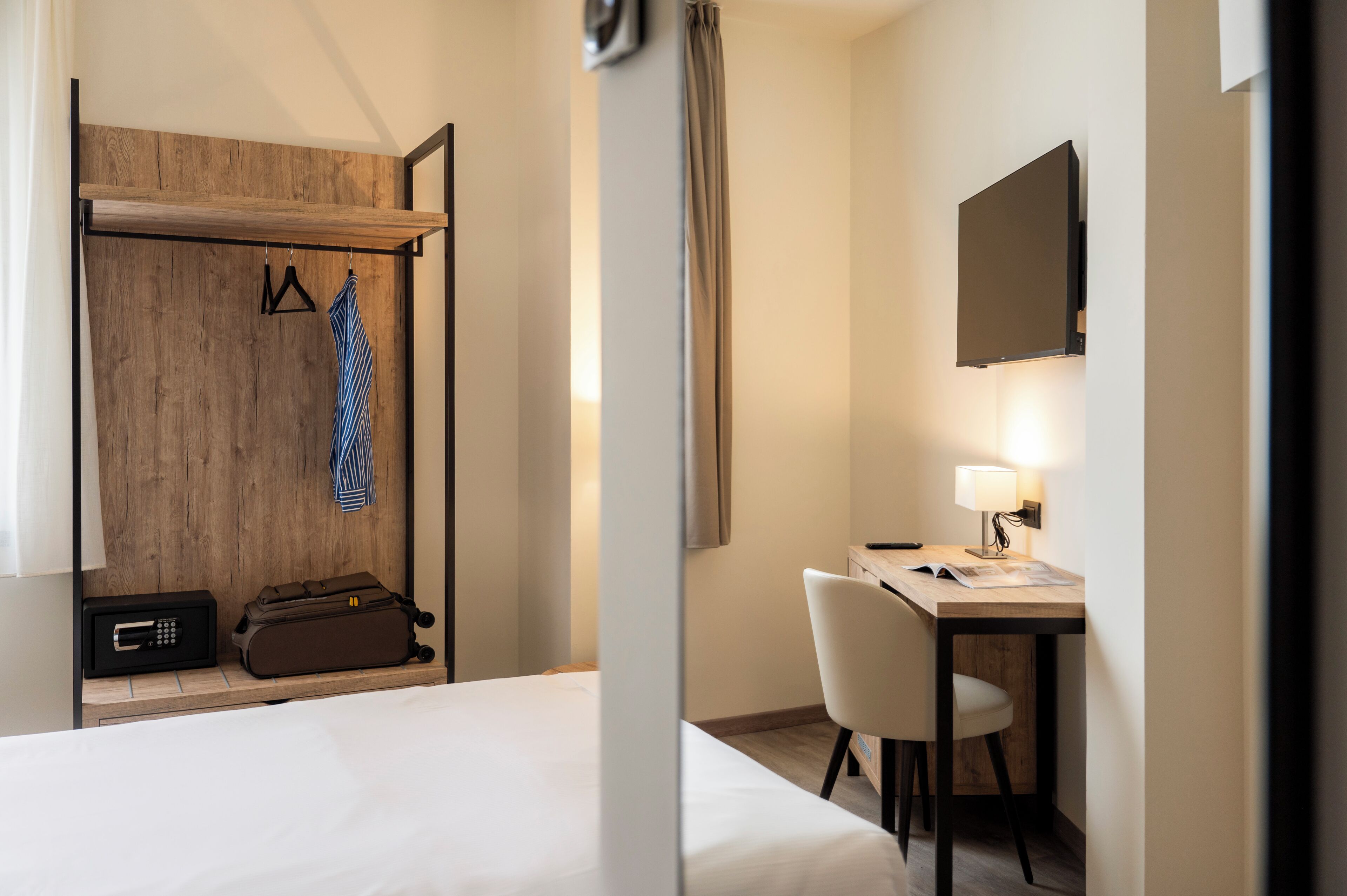 Una habitación de hotel con una cama, un escritorio con silla, un perchero con colgadores, una maleta y un televisor montado en la pared.