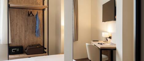 A hotel room with a bed, a desk with a chair, a coat rack with hangers, a suitcase, and a television mounted on the wall.