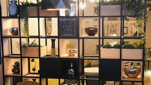 Un cafè acollidor amb taula i cadires de fusta, prestatges amb plantes i objectes decoratius, i un menú a la pissarra.