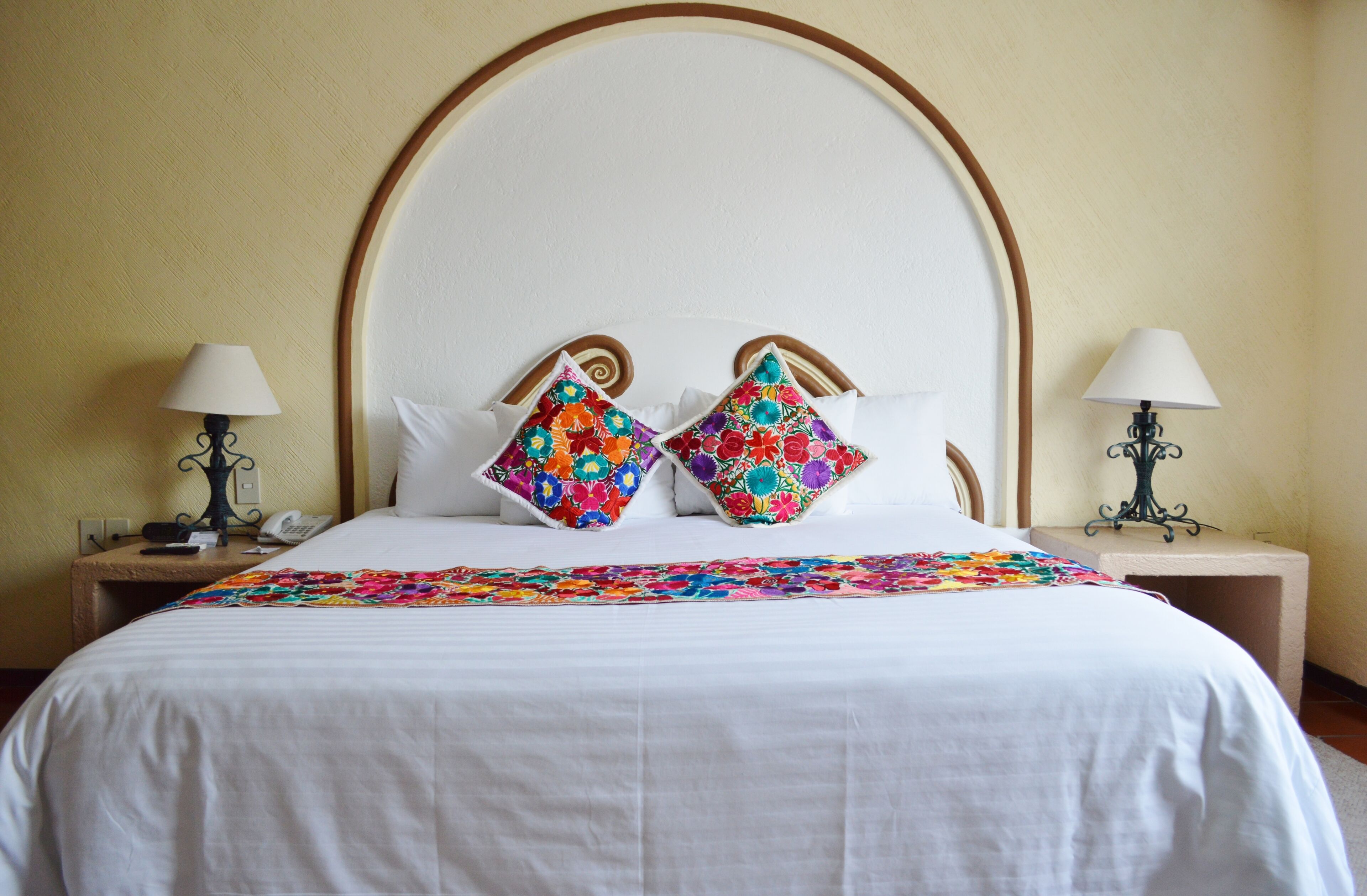 Habitación de hotel con una cama grande, dos almohadones decorativos, una colcha blanca y dos mesitas de noche con lámparas.