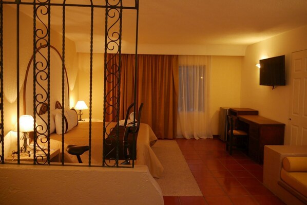 Habitación de hotel con cama, mesita de noche, lámpara, silla, escritorio y televisión en la pared.