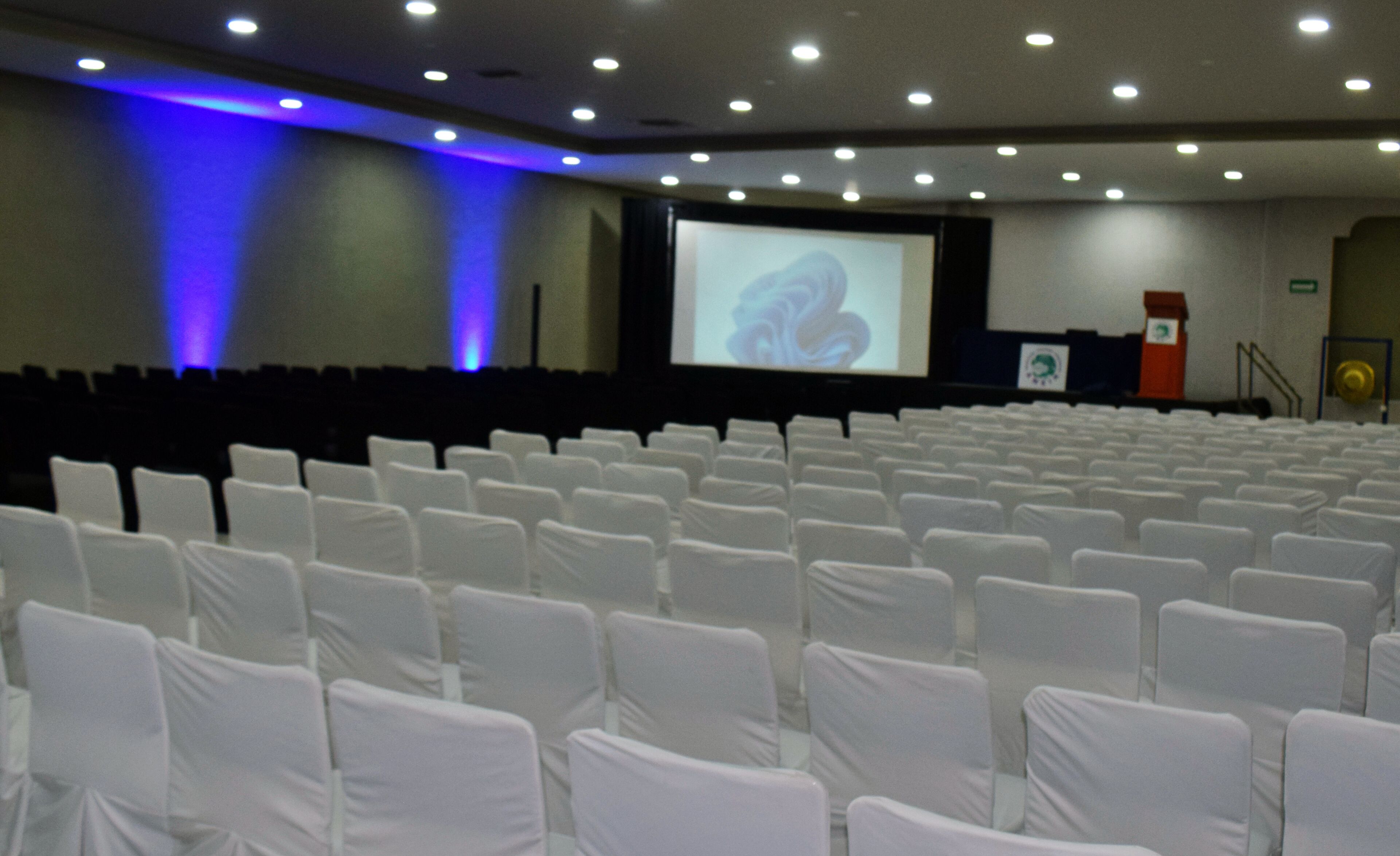 A conference room with rows of white chairs, a large screen, and a podium.