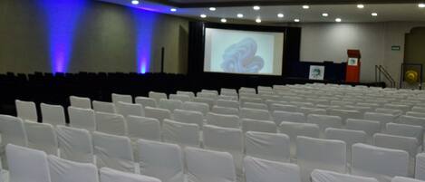 A conference room with rows of white chairs, a large screen, and a podium.