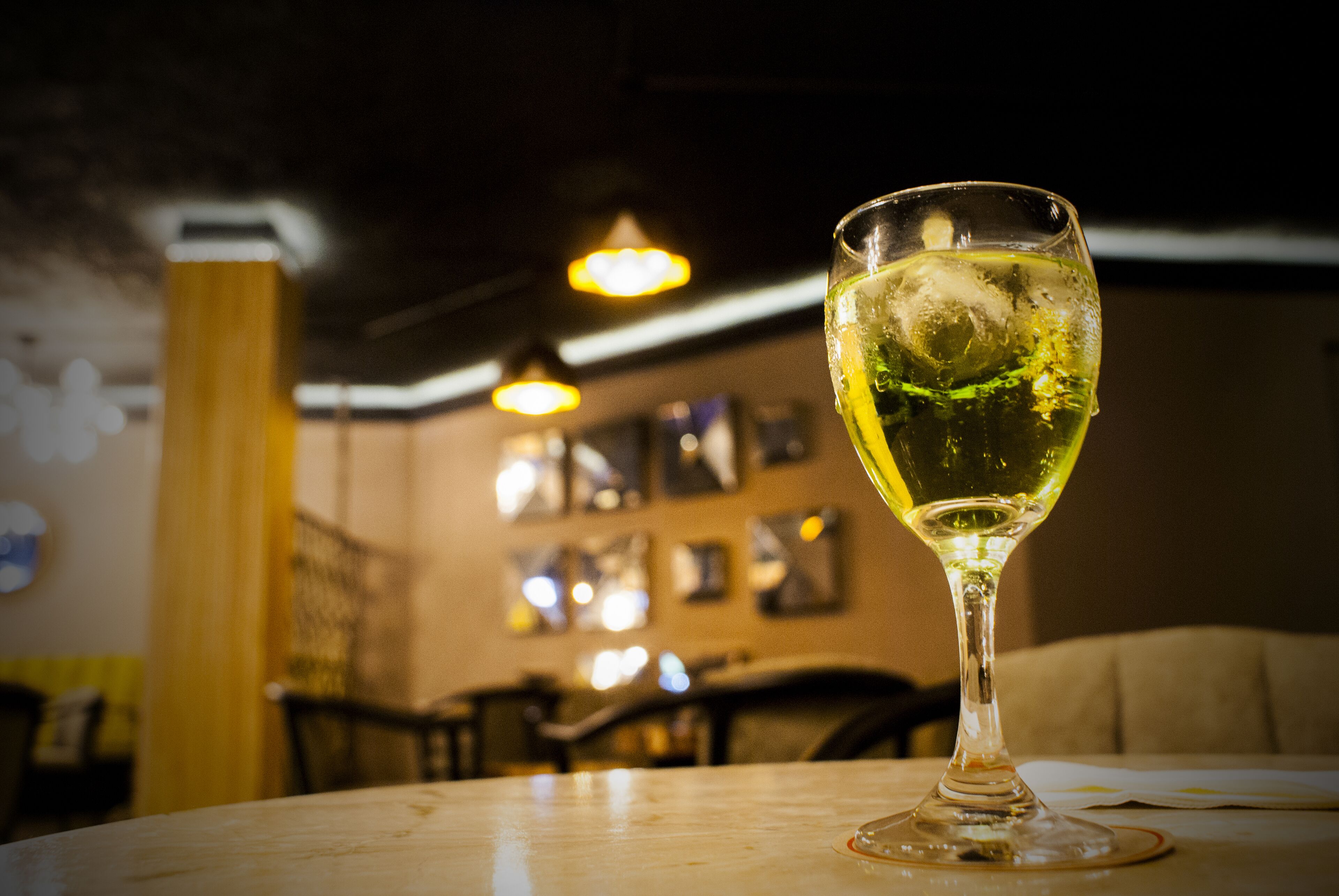 A glass of wine on a table in a dimly lit restaurant with a blurred background of chairs and hanging lights.