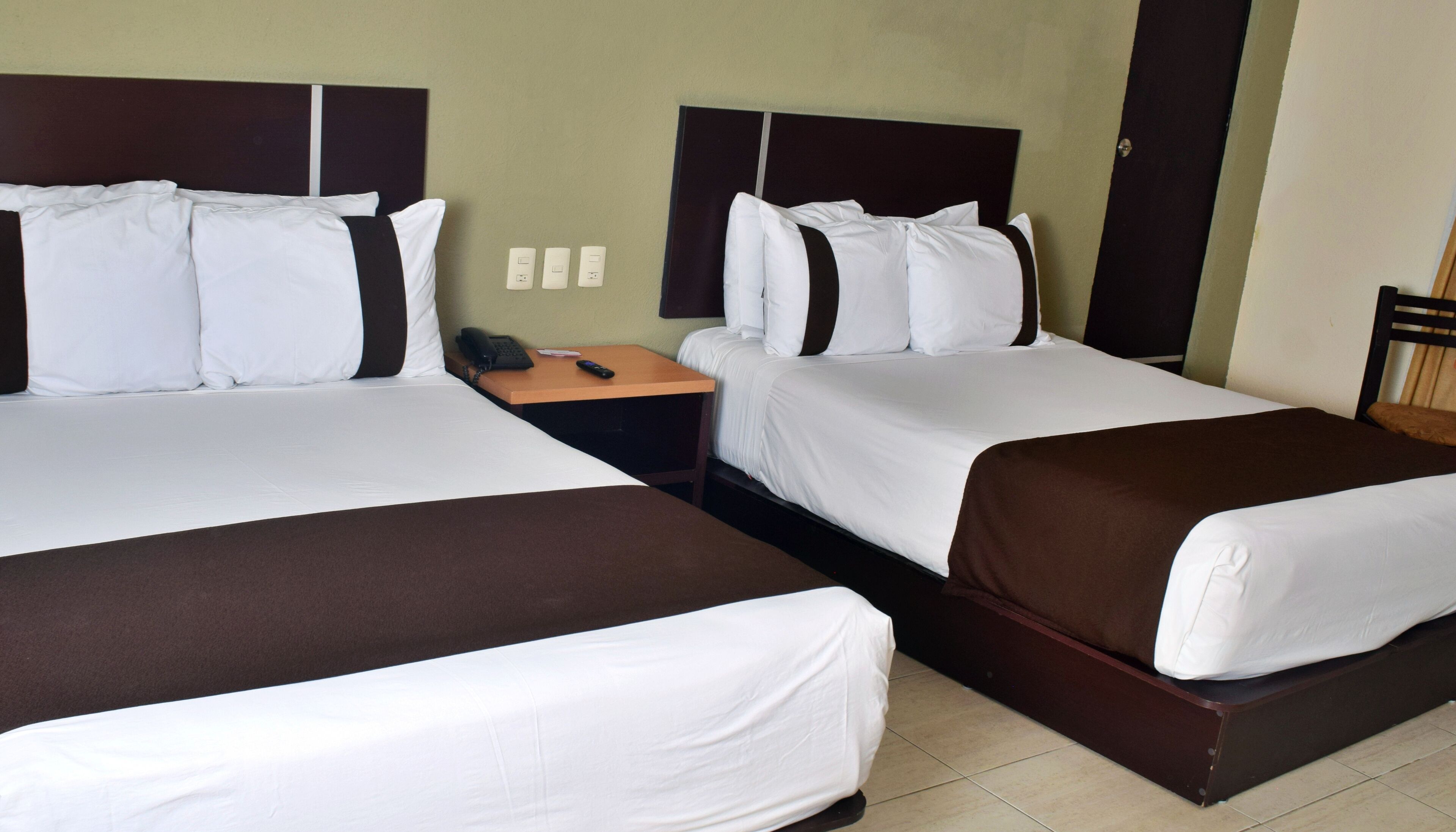 A hotel room with two beds, each with white linens and brown headboards. There is a wooden nightstand with a telephone between the beds.