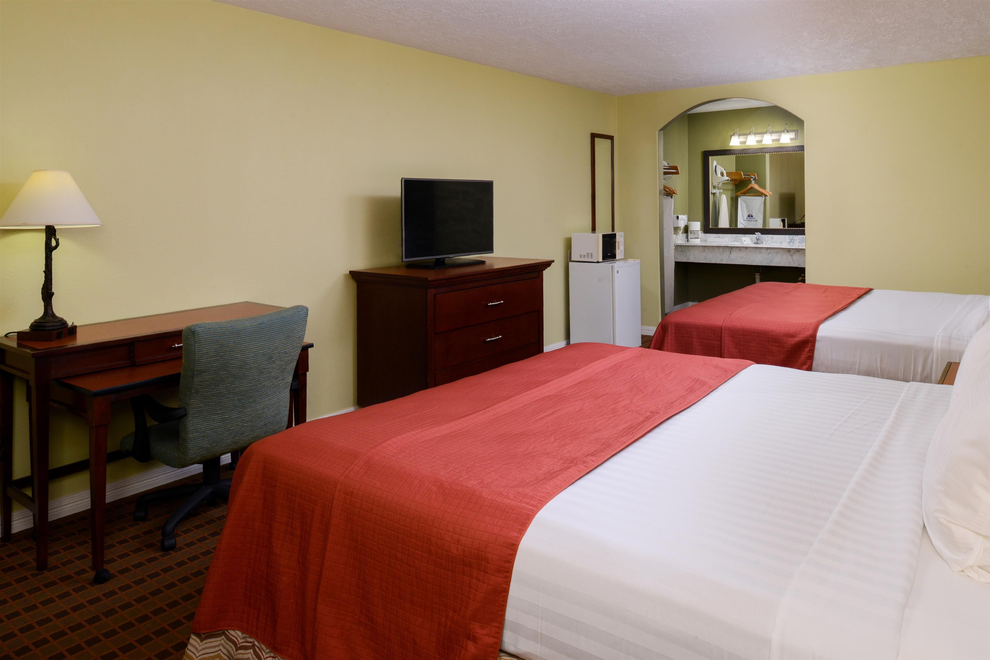 A hotel room with two beds, a desk with a computer, a lamp, a mirror, and a small refrigerator.