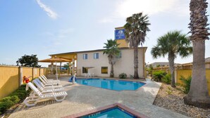 A pool area with lounge chairs, a building with a sign for American Best Value Inn, and palm trees. - Americas Best Value Inn & Suites Waller Prairie View (Waller)