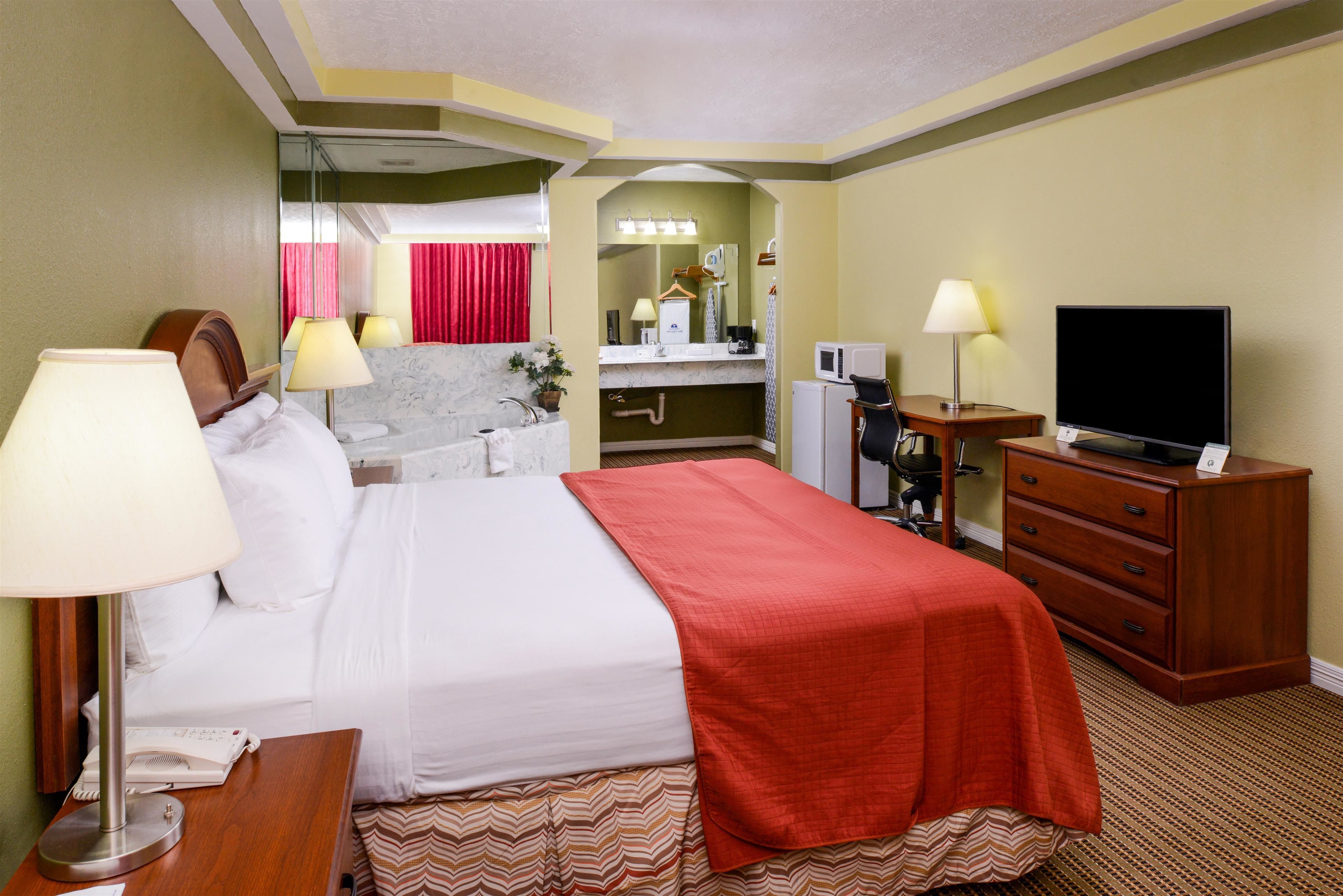 Habitación de hotel con cama, lámparas de noche, televisor, escritorio con lámpara, espejo y baño con lavamanos y ducha.