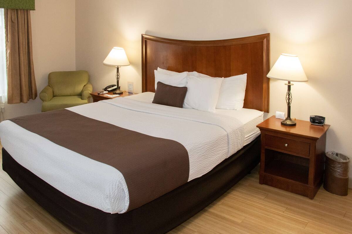 A hotel room with a large bed, two bedside tables with lamps, a green armchair, and a wooden headboard.