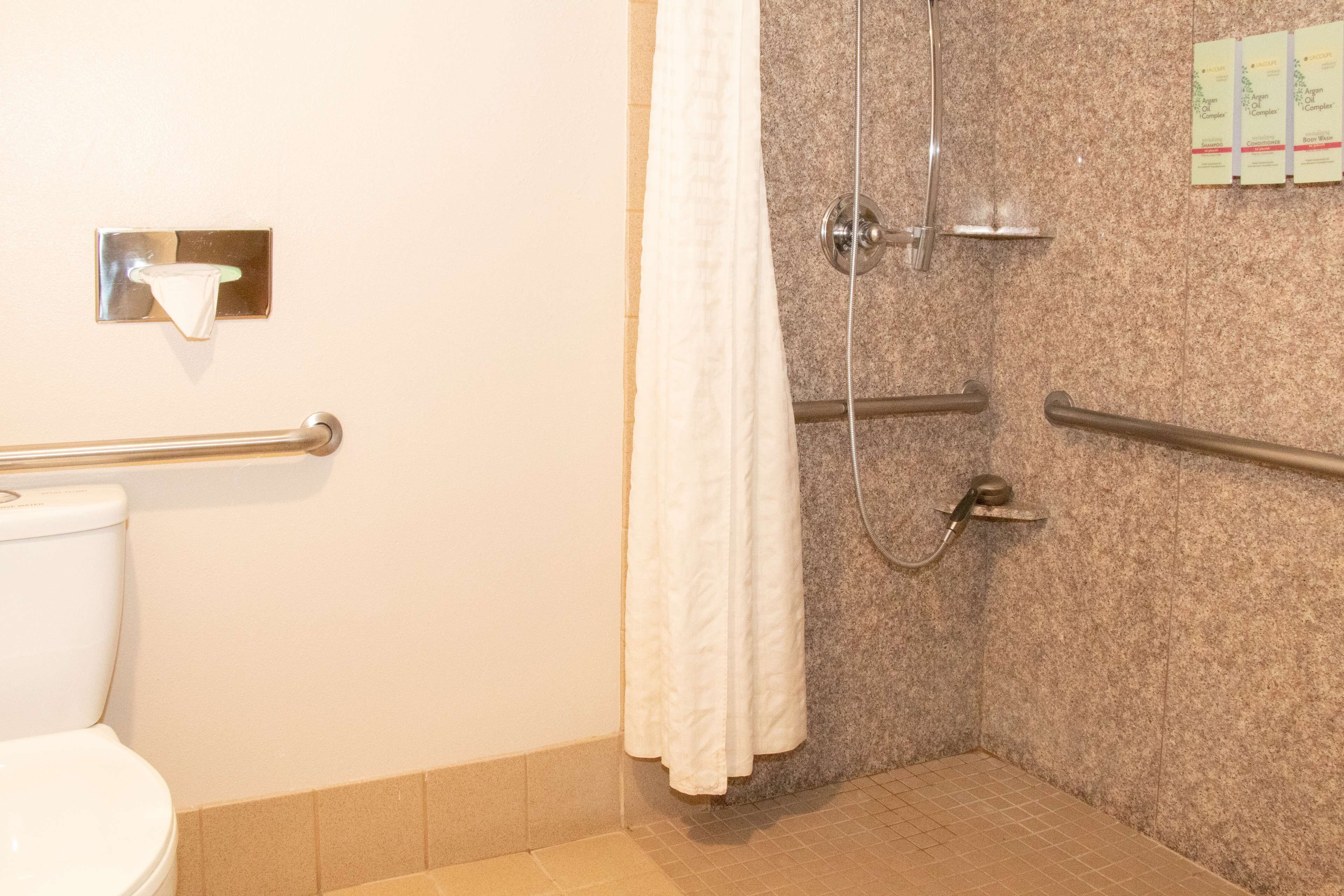 A bathroom with a shower, grab bars, a toilet, and a towel dispenser.