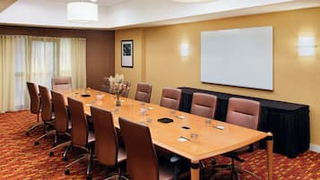 Un salón de conferencias con una larga mesa, sillas marrones, una pantalla de proyector y suelo alfombrado.