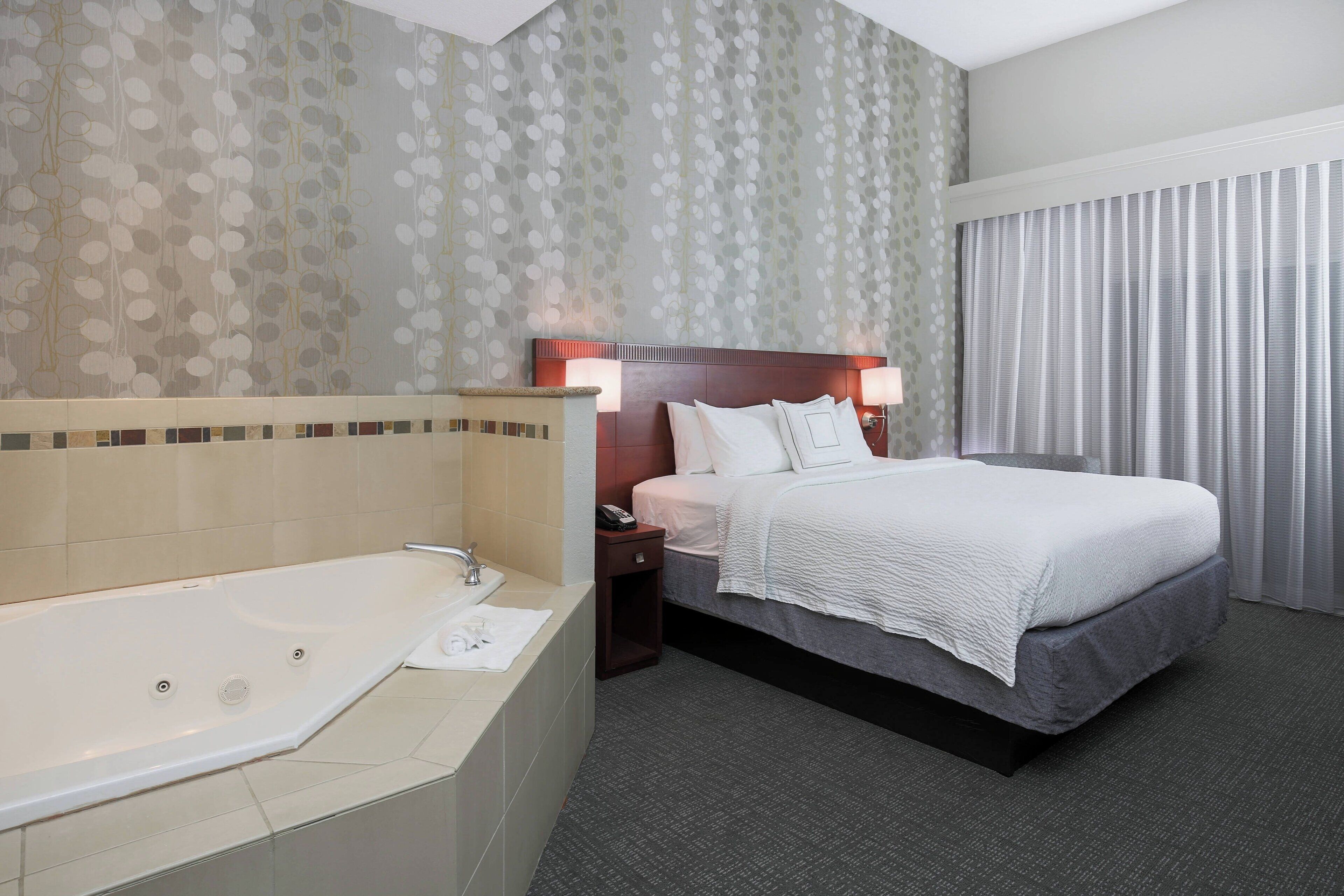 A hotel room with a large bathtub, a bed with white bedding, and a nightstand with a lamp.